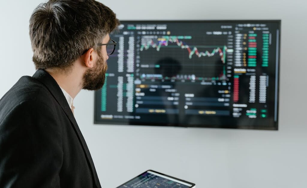 A businessman examines stock market data displayed on a monitor, holding a tablet.